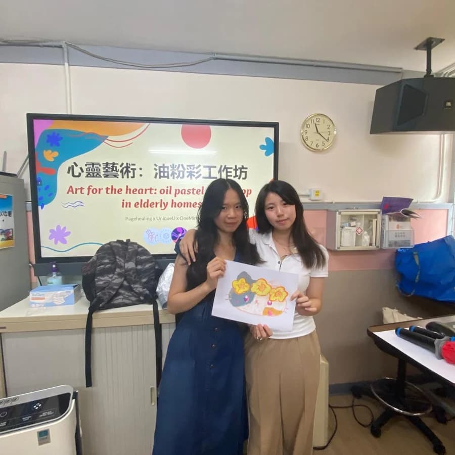 Two volunteers holding up a finished pastel drawing in front of a screen reading 心靈藝術, Art for the heart: oil pastel workshop in elderly homes.