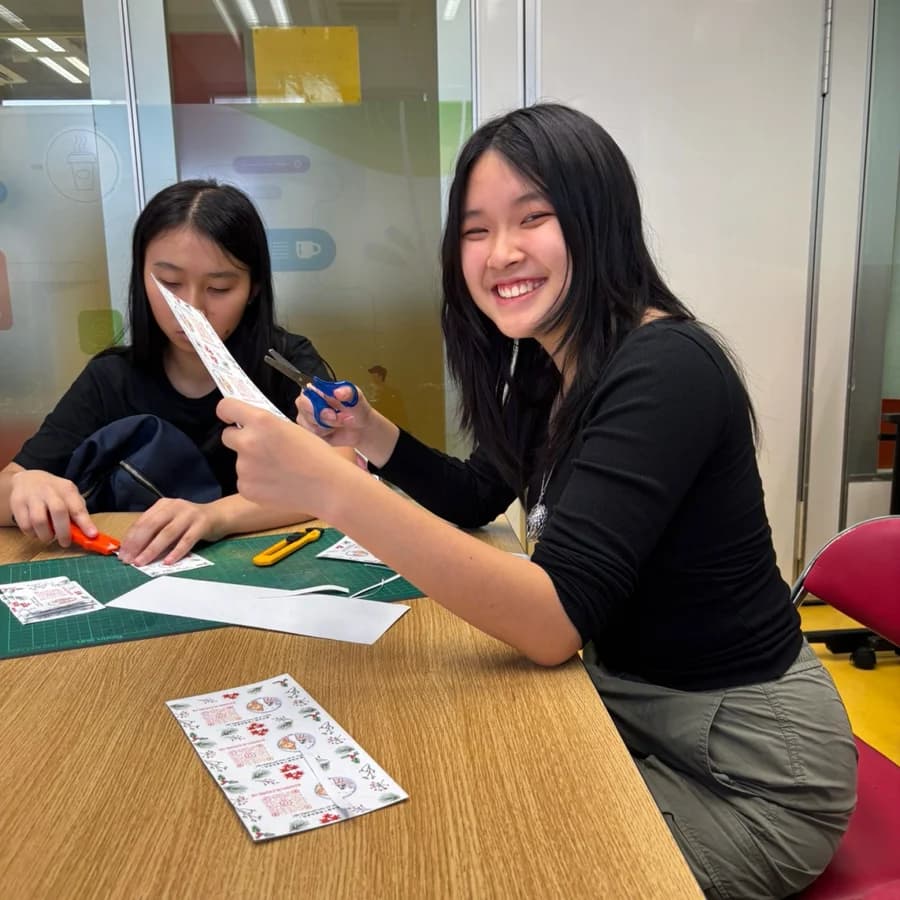 Two volunteers at a table of green cutting mats, scissors and sticker sheets, one holding up a card she has just finished.