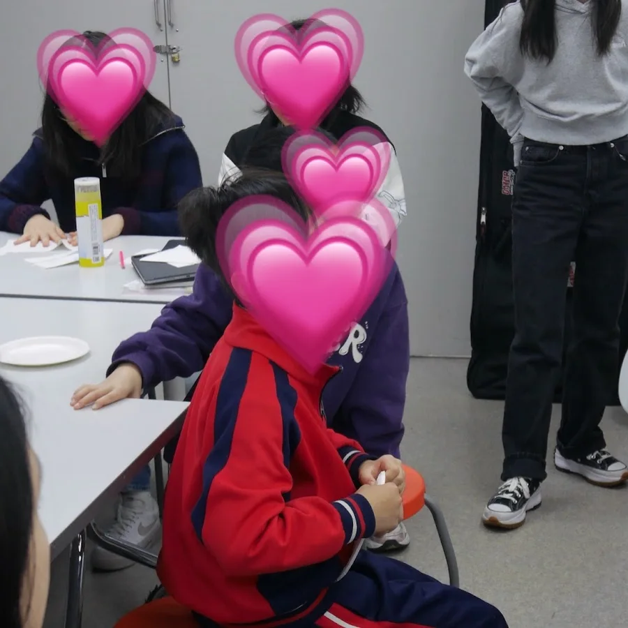 A group standing and sitting around white tables in a bright classroom. Faces are covered with heart stickers.