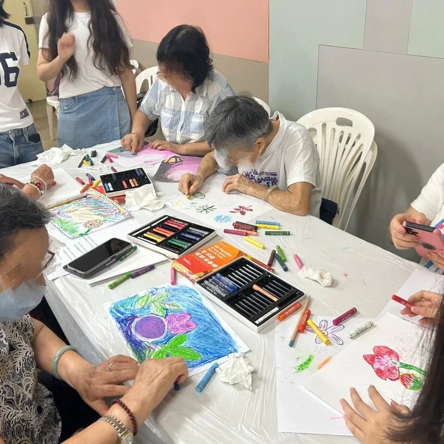 A long table covered in oil pastels and bright finished drawings, with older participants and volunteers working on both sides of it.